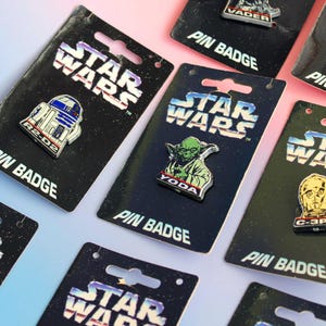 May include: Collection of Star Wars pin badges featuring characters like R2-D2, Yoda, Darth Vader, and C-3PO. Each pin is on a black card with the Star Wars logo and "PIN BADGE" text. The pins have metallic accents.