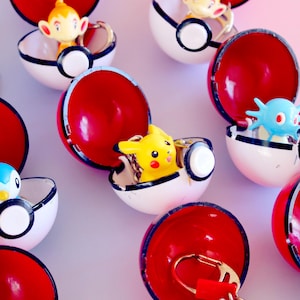 Può includere: Una collezione di portachiavi Poké Ball rossi e bianchi, ognuno con un diverso personaggio Pokémon all'interno. I portachiavi sono aperti, rivelando i personaggi colorati. Lo sfondo è una morbida sfumatura di rosa e bianco.