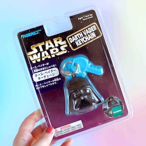 May include: A Star Wars Darth Vader keychain by Tiger Electronics in original blister packaging. The black helmet figure is attached to a blue plastic key-shaped component. The card features the Star Wars logo, text in Japanese, and a Hasbro logo.