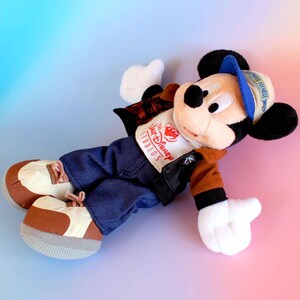May include: A plush Mickey Mouse doll wearing a blue and white cap, a brown and black jacket, blue jeans, and brown and white shoes. The doll has a white shirt with "The Disney Studios" printed on it. The background is a gradient of pink and blue.