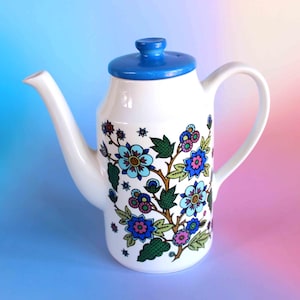 May include: A white ceramic teapot featuring a bright blue lid and a handle on the right side. The body is decorated with a colourful floral pattern consisting of blue, pink, and green flowers with dark green leaves and small star-shaped accents.