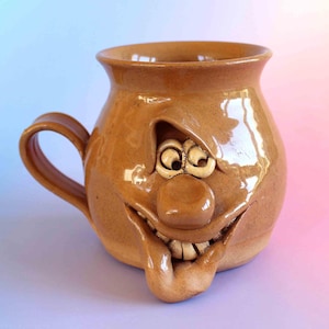Può includere: Una tazza in ceramica color marrone chiaro con un volto scolpito caratterizzato da un naso grande, un ampio sorriso e una lingua sporgente. La tazza presenta una finitura lucida, un corpo arrotondato e un manico curvo sul lato sinistro.
