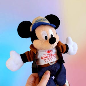 May include: A Mickey Mouse plush toy wearing a brown jacket, blue jeans, and a hat. The white t-shirt has "The Walt Disney Studios" printed on it. The toy has black ears, a black nose, and white gloves.