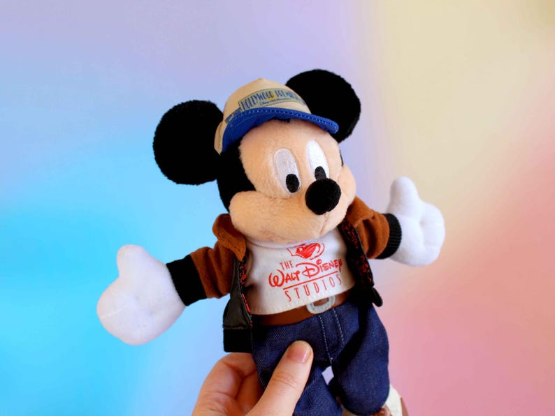 May include: A Mickey Mouse plush toy wearing a brown jacket, blue jeans, and a hat. The white t-shirt has "The Walt Disney Studios" printed on it. The toy has black ears, a black nose, and white gloves.
