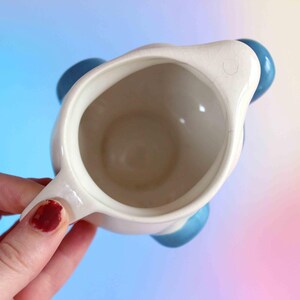 May include: A top-down view of a cream-colored ceramic pitcher with a smooth interior and a small pouring spout. The pitcher features rounded, glossy blue accents on the exterior sides, providing a contrast to the neutral off-white body.