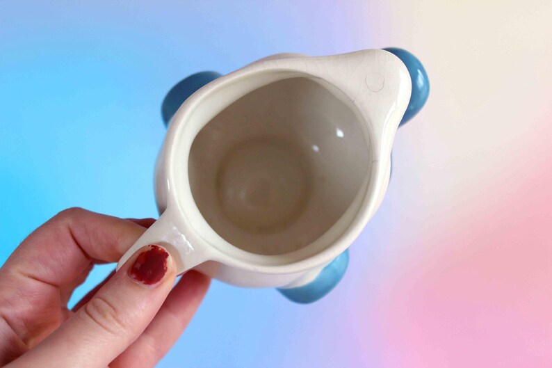 May include: A top-down view of a cream-colored ceramic pitcher with a smooth interior and a small pouring spout. The pitcher features rounded, glossy blue accents on the exterior sides, providing a contrast to the neutral off-white body.