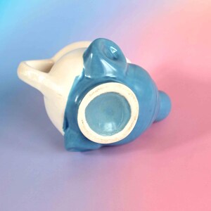 May include: A ceramic pitcher featuring a two-tone design with a glossy cream-colored upper section and a vibrant blue lower section. The vessel includes a curved handle on the side and a circular base, showcasing a smooth, glazed finish throughout.