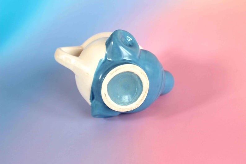 May include: A ceramic pitcher featuring a two-tone design with a glossy cream-colored upper section and a vibrant blue lower section. The vessel includes a curved handle on the side and a circular base, showcasing a smooth, glazed finish throughout.