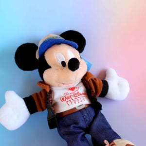 May include: A Mickey Mouse plush toy wearing a brown jacket, blue jeans, and a blue cap. The toy has black ears, white gloves, and a white shirt with "The Walt Disney Studios" printed on it. The background is a gradient of blue and pink.