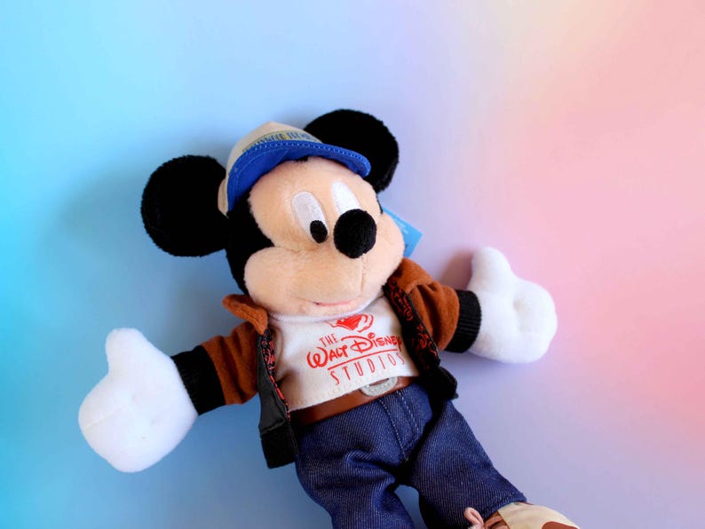 May include: A Mickey Mouse plush toy wearing a brown jacket, blue jeans, and a blue cap. The toy has black ears, white gloves, and a white shirt with "The Walt Disney Studios" printed on it. The background is a gradient of blue and pink.