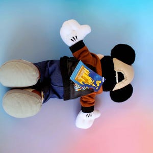 May include: A Mickey Mouse plush toy wearing a brown jacket, blue jeans, and white gloves and shoes. The plush has black ears and a tan face. A tag is attached to the jacket.