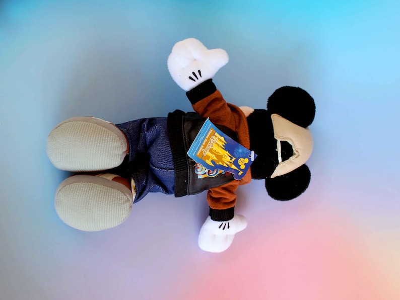 May include: A Mickey Mouse plush toy wearing a brown jacket, blue jeans, and white gloves and shoes. The plush has black ears and a tan face. A tag is attached to the jacket.