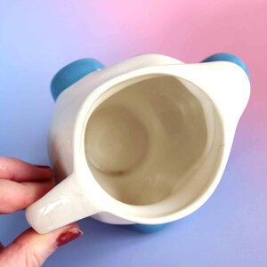 May include: A top-down view of a white ceramic pitcher with a smooth interior and a handle on the side. The pitcher features rounded blue accents on the exterior rim, visible from this overhead perspective, showcasing a simple and functional design.