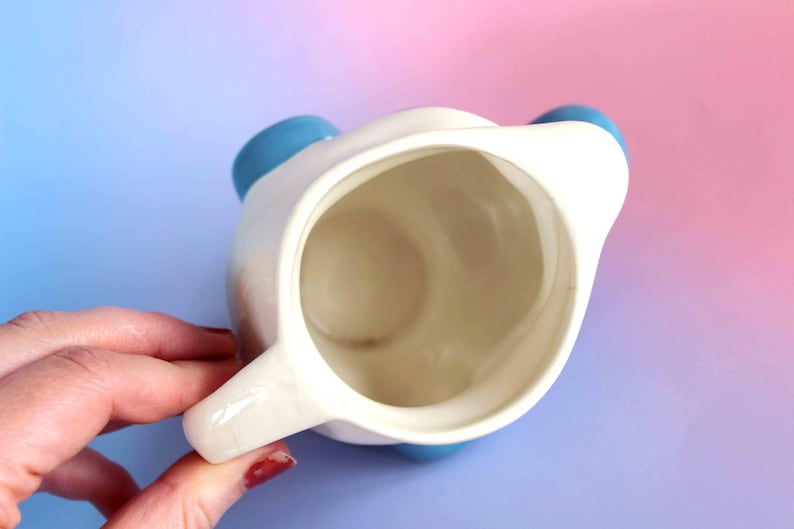 May include: A top-down view of a white ceramic pitcher with a smooth interior and a handle on the side. The pitcher features rounded blue accents on the exterior rim, visible from this overhead perspective, showcasing a simple and functional design.