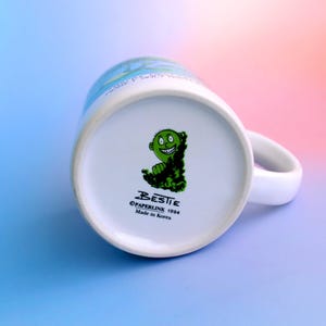 K&ouml;nnte beinhalten: Wei&szlig;er Keramikbecher mit gebogenem Henkel. Der Boden des Bechers zeigt eine gr&uuml;ne Cartoonfigur und den Text "BESTIE &copy;PAPERLINE 1994 Made in Korea". Der Becher steht vor einem Farbverlauf aus Blau, Rosa und Lila.