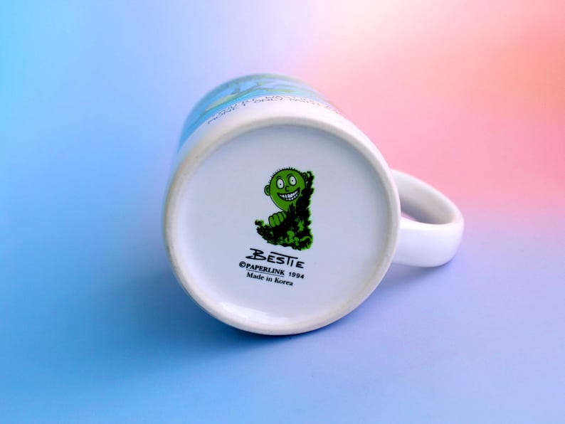 K&ouml;nnte beinhalten: Wei&szlig;er Keramikbecher mit gebogenem Henkel. Der Boden des Bechers zeigt eine gr&uuml;ne Cartoonfigur und den Text "BESTIE &copy;PAPERLINE 1994 Made in Korea". Der Becher steht vor einem Farbverlauf aus Blau, Rosa und Lila.
