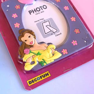 May include: A purple oval photo frame featuring a cartoon character in a yellow dress holding a pink rose. The frame is decorated with small pink flowers and sparkles. The packaging includes the text PHOTO, FOTO, BILD, Hang Tag, Stand, and DECOFUN.