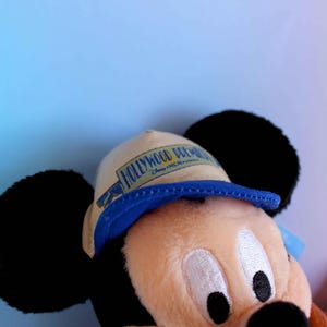 May include: A plush Mickey Mouse toy wearing a tan and blue hat with the text "HOLLYWOOD DREAM" printed on it. The toy has black ears, a tan face, and a black nose. The background is a gradient of blue and pink.