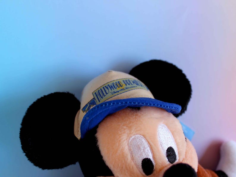 May include: A plush Mickey Mouse toy wearing a tan and blue hat with the text "HOLLYWOOD DREAM" printed on it. The toy has black ears, a tan face, and a black nose. The background is a gradient of blue and pink.