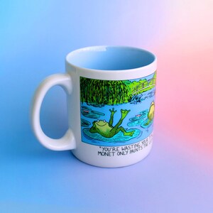 K&ouml;nnte beinhalten: Wei&szlig;er Keramikbecher mit hellblauem Innenraum und wei&szlig;em Henkel. Der Becher zeigt eine Cartoon-Illustration eines Frosches, der sich auf einem Seerosenblatt entspannt, mit dem Text "YOU'RE WASTING YOUR MONET ONLY PAINTS THE..." an der Seite.