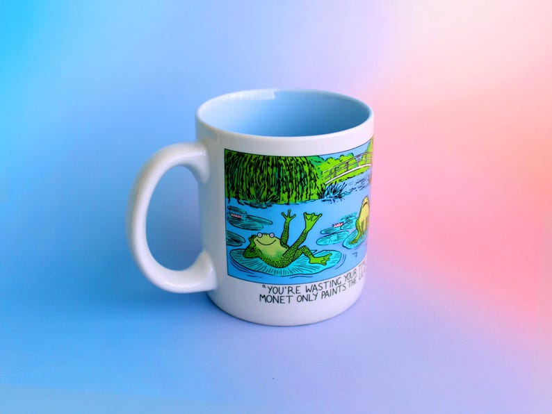 K&ouml;nnte beinhalten: Wei&szlig;er Keramikbecher mit hellblauem Innenraum und wei&szlig;em Henkel. Der Becher zeigt eine Cartoon-Illustration eines Frosches, der sich auf einem Seerosenblatt entspannt, mit dem Text "YOU'RE WASTING YOUR MONET ONLY PAINTS THE..." an der Seite.