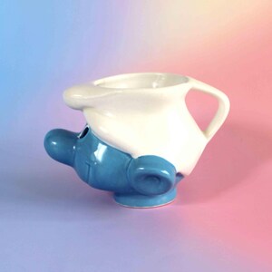 May include: A ceramic mug shaped like a blue character head wearing a white hat. The mug features a prominent blue nose, a small ear, and a white handle integrated into the hat design. The surface is glossy, showing a blue face and white cap.