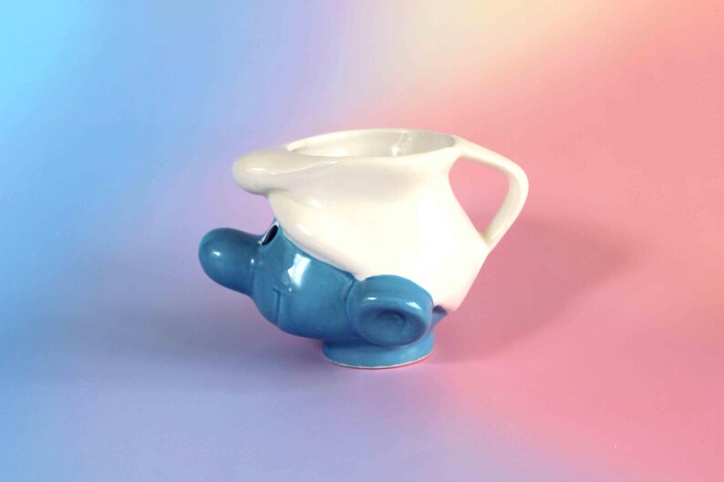 May include: A ceramic mug shaped like a blue character head wearing a white hat. The mug features a prominent blue nose, a small ear, and a white handle integrated into the hat design. The surface is glossy, showing a blue face and white cap.