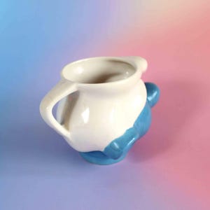 May include: A ceramic pitcher featuring a two-tone design with a glossy white upper section and a vibrant blue lower base. The vessel includes a curved handle on the side and a wide, rounded opening at the top, showcasing a smooth, reflective finish.