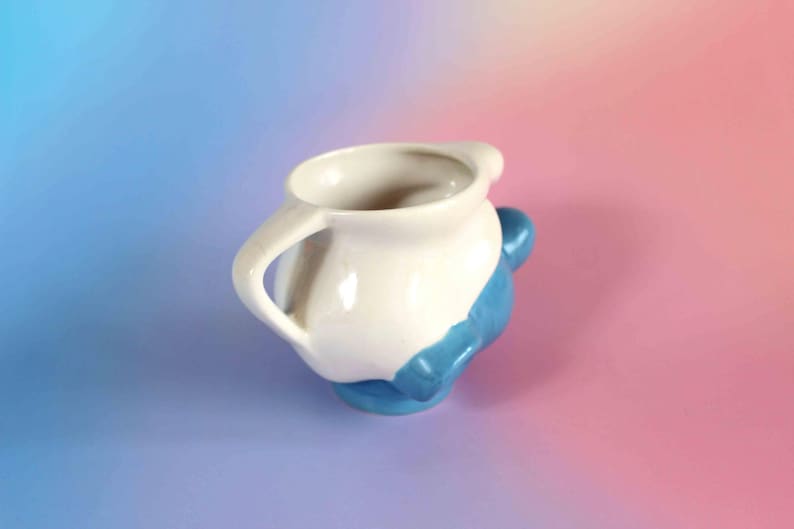 May include: A ceramic pitcher featuring a two-tone design with a glossy white upper section and a vibrant blue lower base. The vessel includes a curved handle on the side and a wide, rounded opening at the top, showcasing a smooth, reflective finish.