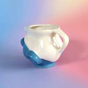 May include: A ceramic mug featuring a two-tone design with a glossy white upper section and a solid blue lower base. The mug has a rounded, organic shape with a single handle on the right side and several small, rounded blue protrusions on the exterior.