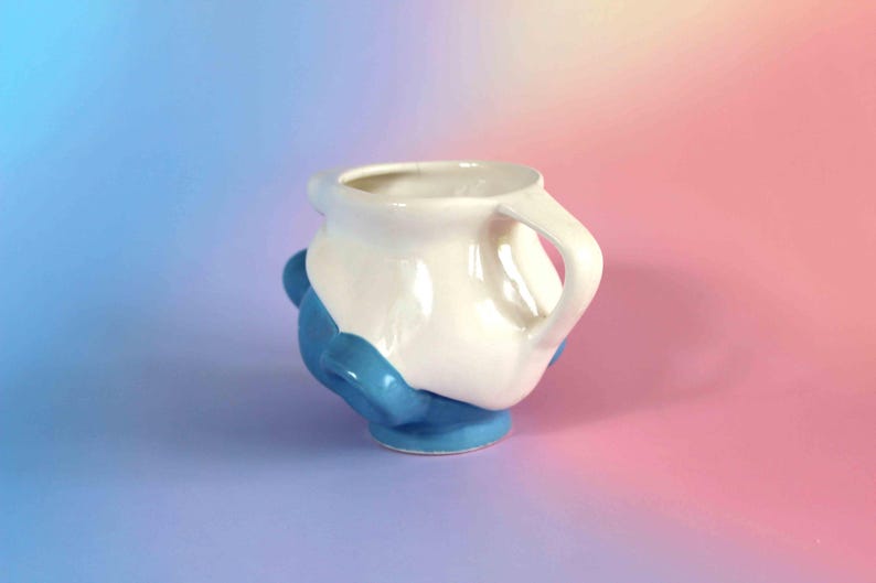 May include: A ceramic mug featuring a two-tone design with a glossy white upper section and a solid blue lower base. The mug has a rounded, organic shape with a single handle on the right side and several small, rounded blue protrusions on the exterior.