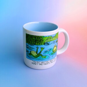 K&ouml;nnte beinhalten: Wei&szlig;er Keramikbecher mit hellblauem Innenraum und Henkel. Der Becher zeigt eine Cartoon-Illustration von Fr&ouml;schen in einem Teich mit dem Text "YOU'RE WASTING YOUR TIME MONET ONLY PAINTS THE LILIES."
