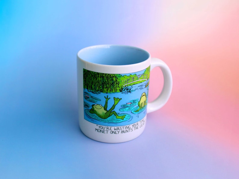 K&ouml;nnte beinhalten: Wei&szlig;er Keramikbecher mit hellblauem Innenraum und Henkel. Der Becher zeigt eine Cartoon-Illustration von Fr&ouml;schen in einem Teich mit dem Text "YOU'RE WASTING YOUR TIME MONET ONLY PAINTS THE LILIES."