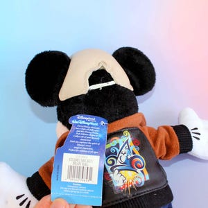 May include: A Mickey Mouse Studio bean bag plush toy. The plush has black ears, a tan hat, and white gloves. It wears a brown jacket with a colorful graphic on the back. A blue tag reads "Disneyland" and "Studio Mickey Bean Bag".