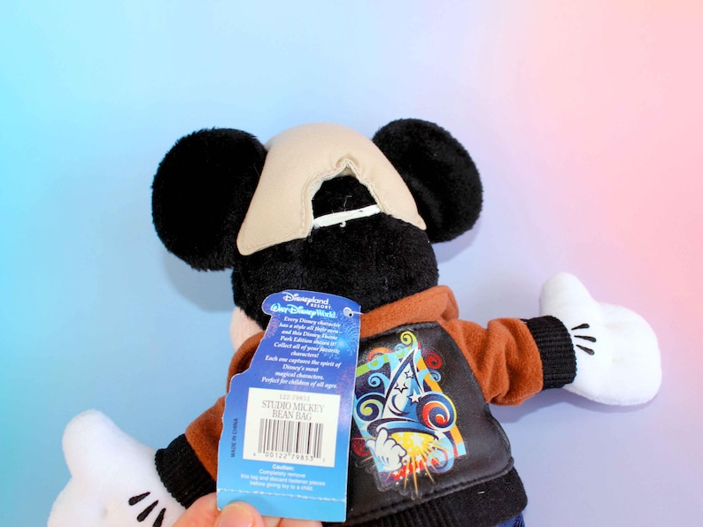 May include: A Mickey Mouse Studio bean bag plush toy. The plush has black ears, a tan hat, and white gloves. It wears a brown jacket with a colorful graphic on the back. A blue tag reads "Disneyland" and "Studio Mickey Bean Bag".