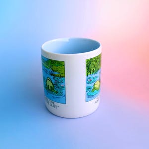 K&ouml;nnte beinhalten: Wei&szlig;er Keramikbecher mit hellblauem Innenraum. Der Becher zeigt eine Cartoon-Illustration eines Frosches in einem Teich mit dem Text "Bestie" und "More Time for the Lilies". Der Becher steht vor einem Farbverlauf aus Blau und Rosa.