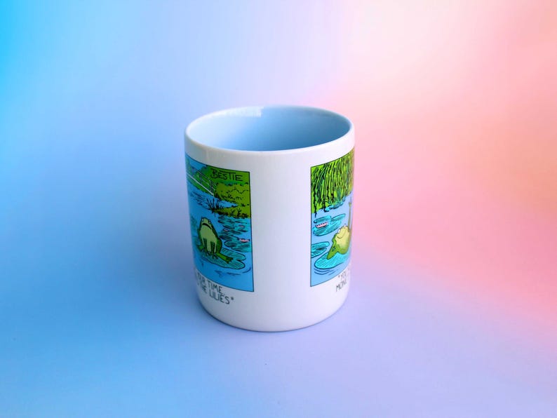 K&ouml;nnte beinhalten: Wei&szlig;er Keramikbecher mit hellblauem Innenraum. Der Becher zeigt eine Cartoon-Illustration eines Frosches in einem Teich mit dem Text "Bestie" und "More Time for the Lilies". Der Becher steht vor einem Farbverlauf aus Blau und Rosa.