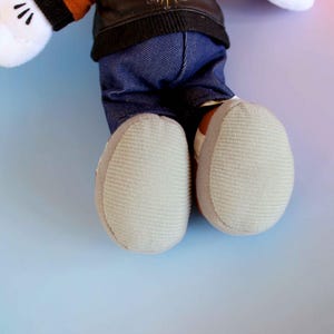 May include: A plush toy featuring white gloves with black details, a black jacket, blue denim pants, and light gray shoes. The toy's feet are visible, showcasing the textured soles. The background is a gradient of blue, pink, and purple.