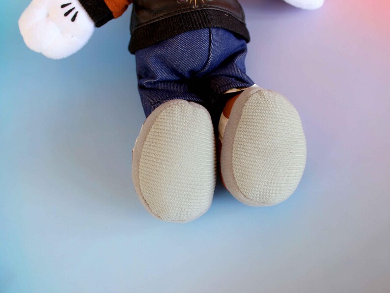 May include: A plush toy featuring white gloves with black details, a black jacket, blue denim pants, and light gray shoes. The toy's feet are visible, showcasing the textured soles. The background is a gradient of blue, pink, and purple.