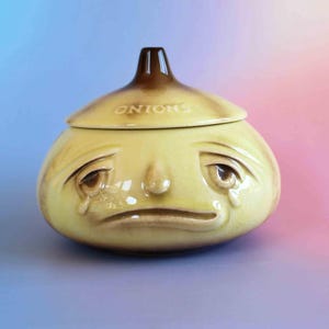 May include: A ceramic onion-shaped storage jar with a glossy light yellow finish and brown accents. The lid features the word ONIONS embossed on the top. The jar has a sculpted face with sad, drooping eyes, tear-like markings, and a downturned mouth.