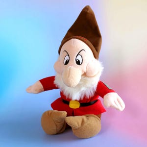 Peut inclure: Une peluche représentant le personnage Grincheux de Blanche-Neige, avec un bonnet pointu marron, une tunique rouge, une ceinture noire à boucle jaune, des chaussures beiges et une barbe en fausse fourrure blanche. Il a un air renfrogné.
