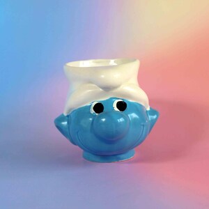 May include: A ceramic mug shaped like a blue Smurf character wearing a white hat. The mug features a bright blue face with a large round nose, black circular eyes, and a wide smiling mouth, topped with a white hat that forms the rim of the mug.