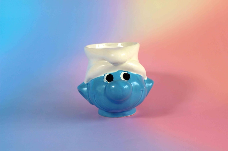May include: A ceramic mug shaped like a blue Smurf character wearing a white hat. The mug features a bright blue face with a large round nose, black circular eyes, and a wide smiling mouth, topped with a white hat that forms the rim of the mug.