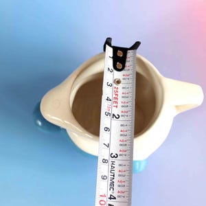 May include: A cream-colored ceramic pitcher with a blue handle and base, viewed from above. A white measuring tape is placed across the opening, showing a diameter of approximately 3.5 inches. The pitcher has a smooth, rounded body.