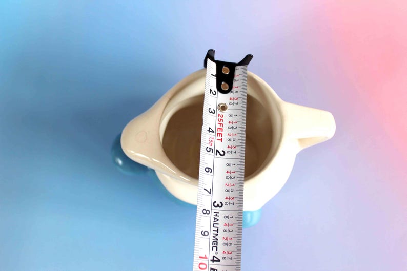 May include: A cream-colored ceramic pitcher with a blue handle and base, viewed from above. A white measuring tape is placed across the opening, showing a diameter of approximately 3.5 inches. The pitcher has a smooth, rounded body.