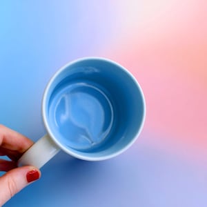 K&ouml;nnte beinhalten: Eine hellblaue Keramik-Tasse mit wei&szlig;em Henkel. Das Innere der Tasse ist ein hellerer Blauton. Der Hintergrund ist ein Farbverlauf aus Blau, Rosa und Orange.