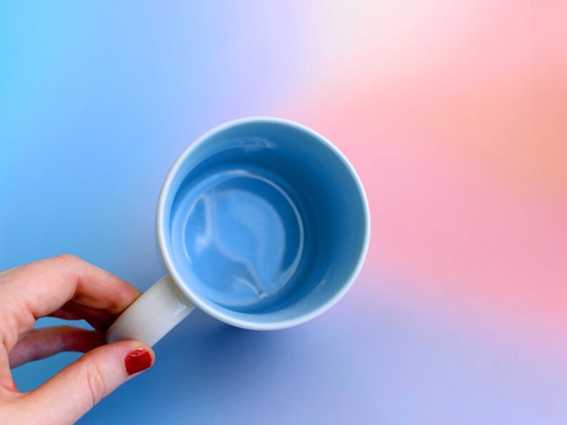 K&ouml;nnte beinhalten: Eine hellblaue Keramik-Tasse mit wei&szlig;em Henkel. Das Innere der Tasse ist ein hellerer Blauton. Der Hintergrund ist ein Farbverlauf aus Blau, Rosa und Orange.