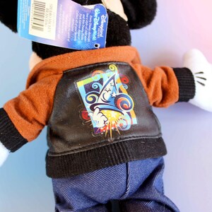May include: A Mickey Mouse plush toy wearing a brown and black jacket with a colorful graphic on the back. The jacket has a hood and is paired with blue denim pants. The toy has white gloves and black ears.