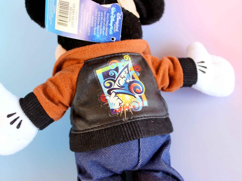 May include: A Mickey Mouse plush toy wearing a brown and black jacket with a colorful graphic on the back. The jacket has a hood and is paired with blue denim pants. The toy has white gloves and black ears.