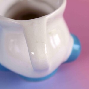 May include: A white ceramic pitcher featuring a glossy finish with visible fine crackle patterns throughout the glaze. The pitcher has a curved handle and is supported by three rounded, bright blue feet positioned at the base of the vessel.